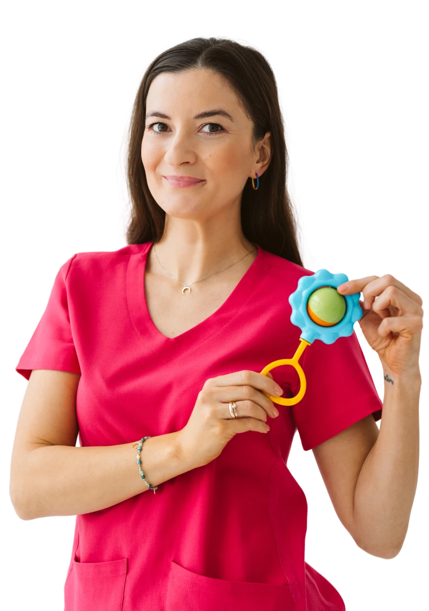 Izabella Malara-Zygaj w różowym uniformie medycznym, uśmiecha się i trzyma kolorową zabawkę sensoryczną dla niemowląt.